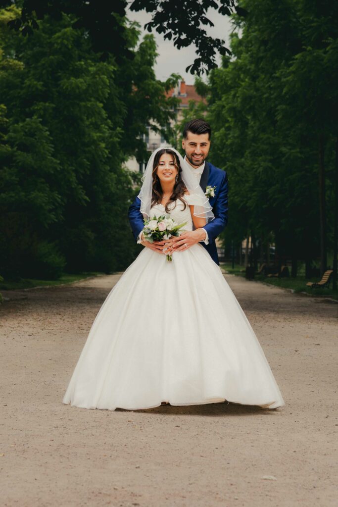 Marié tenant sa femme devant lui dans une allée de parc, avec son bouquet