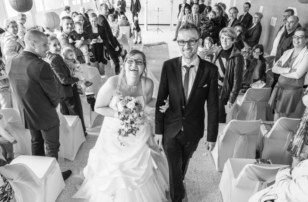 Mariés sortant d'une cérémonie dans une allée, face à l'objectif, entourés de leurs invités