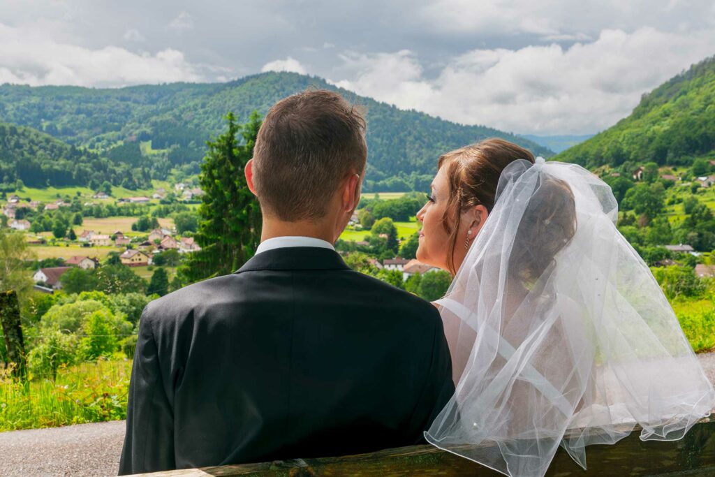 Couple de mariés assis sur un banc face à un paysage de montagnes et de forêt