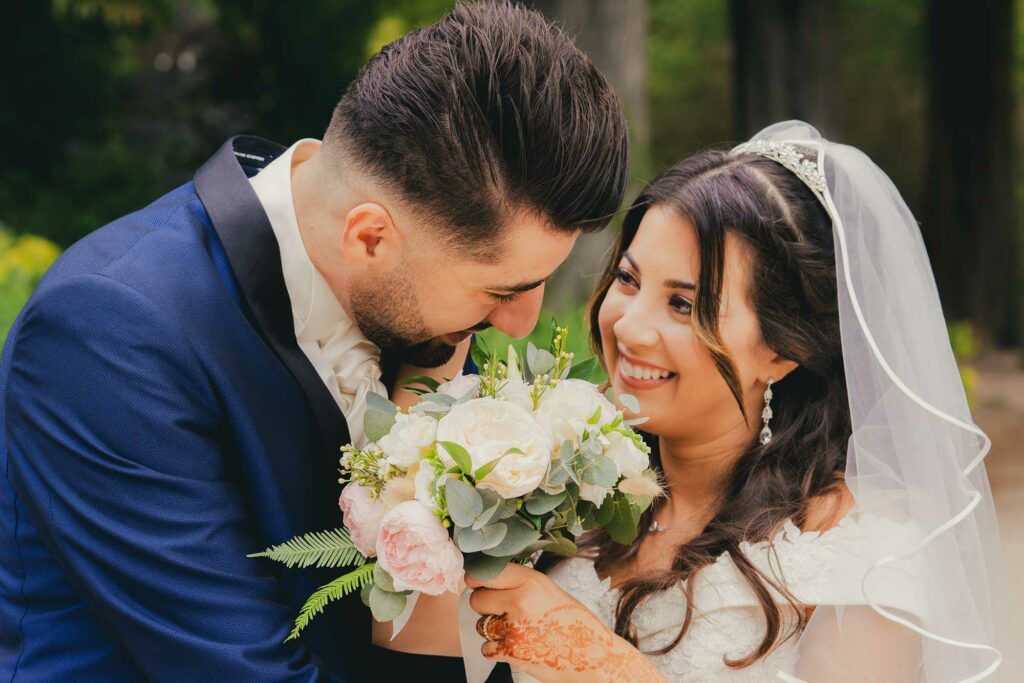 Mariée touchant le visage de son marié, bouquet de fleurs à la main, dans un parc